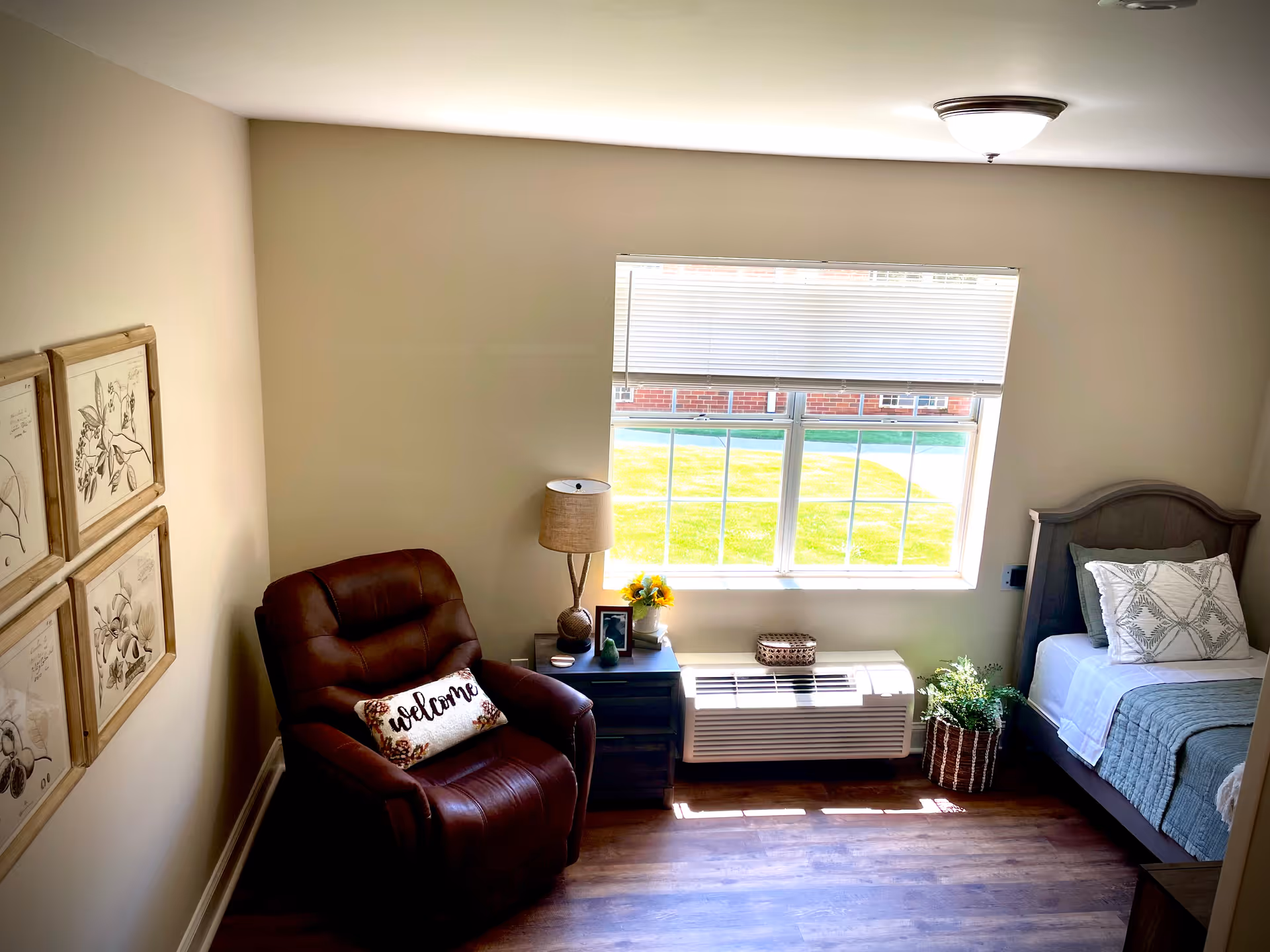 A cozy bedroom with a single bed on the right side, a bedside table with a lamp and decorative items, a large window with blinds partially open letting in sunlight, a comfortable brown recliner chair with a 'welcome' pillow, and framed botanical artwork on the left wall.