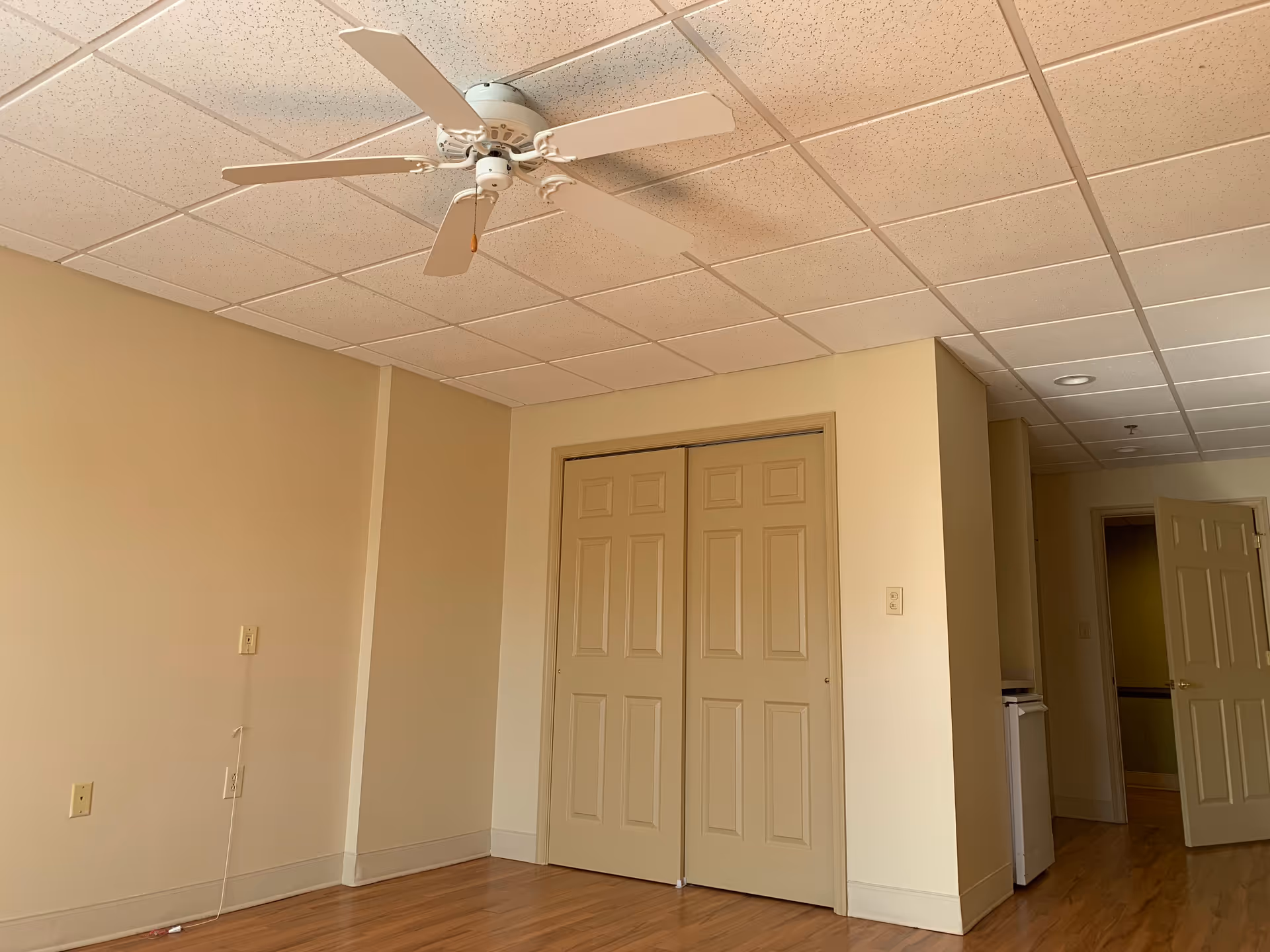 Empty room with beige walls, wooden floor, a white ceiling fan, and a double-door closet. There is an open door leading to another room and a small white appliance partially visible in the hallway.