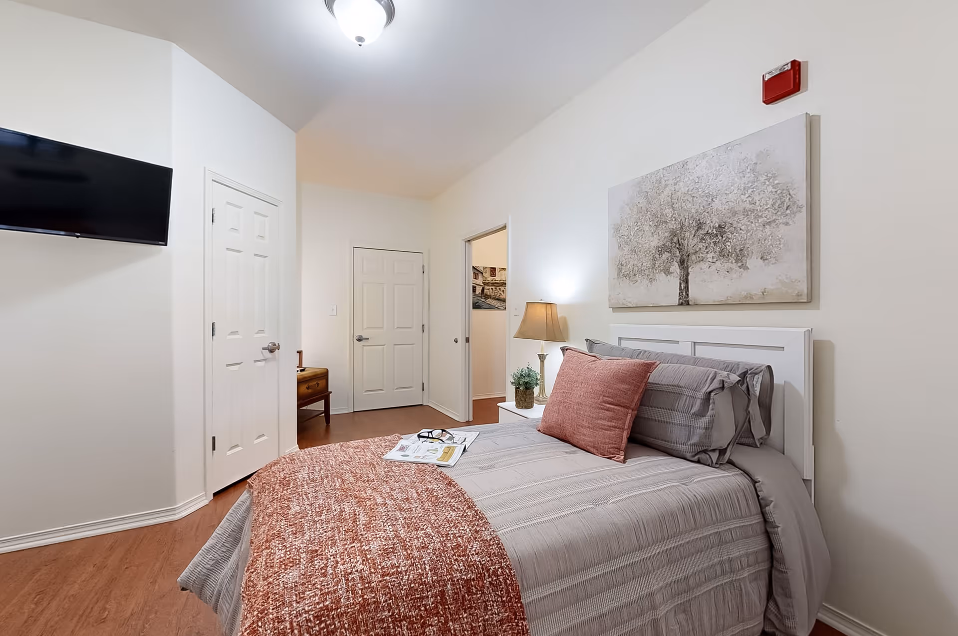 A cozy bedroom with a neatly made bed featuring gray and rust-colored bedding. A nightstand with a lamp and a small plant is beside the bed. Above the bed hangs a painting of a tree. There is a wall-mounted TV on the left, two closed doors, and a small wooden table in the corner. The room has light-colored walls and wooden flooring.