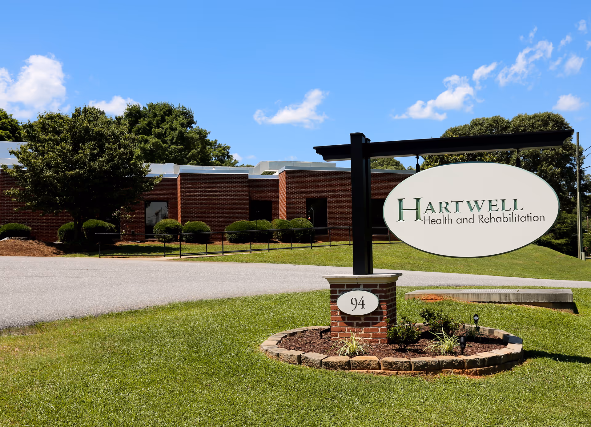 Exterior view of Hartwell Health and Rehabilitation building with a large sign displaying the facility name and the number 94 on a brick post, surrounded by green grass, bushes, and trees under a blue sky with some clouds.