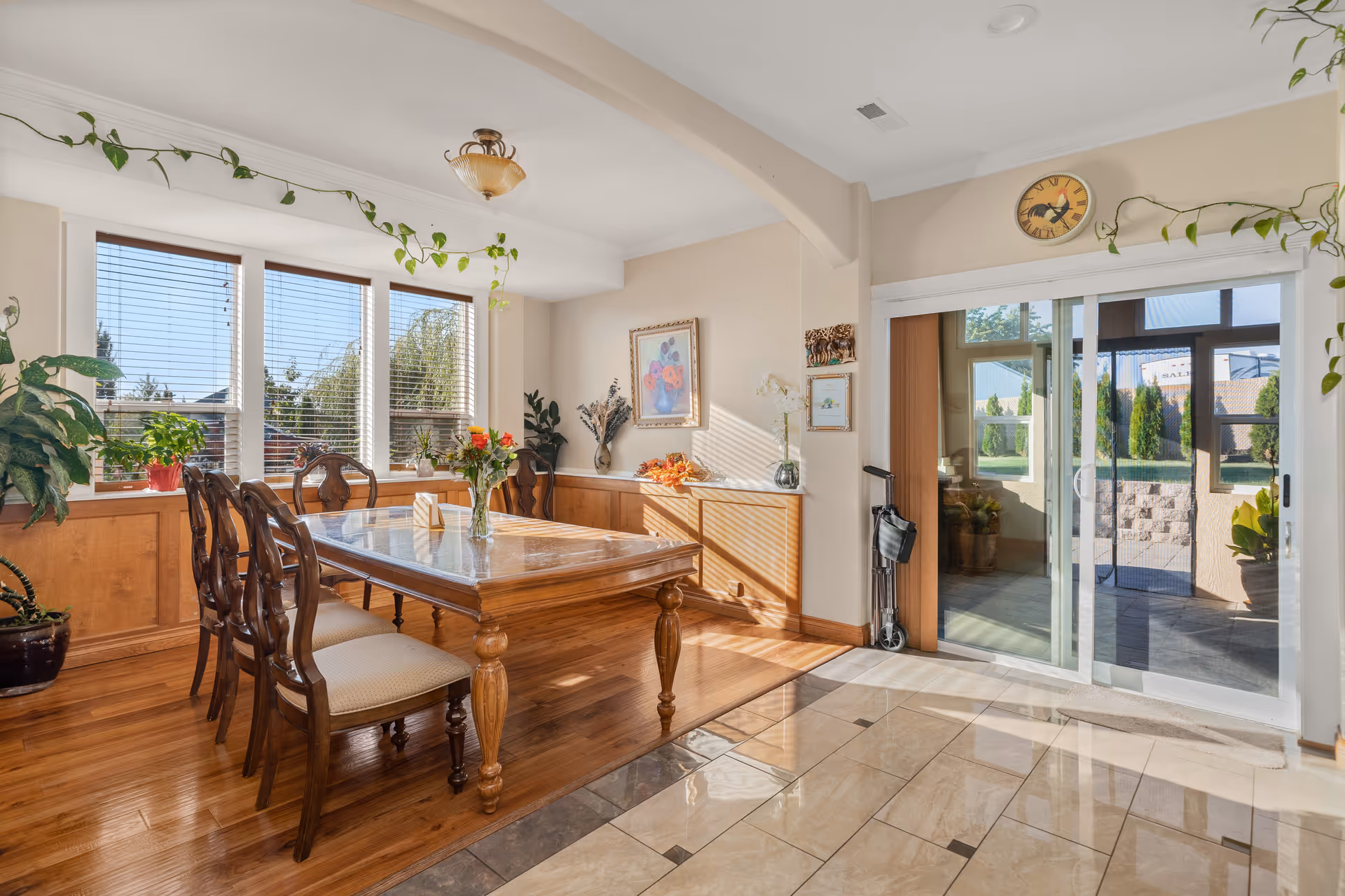 A bright dining room with a wooden table and six chairs. The room features large windows with blinds, letting in natural light. There are plants on the windowsill and a vase with flowers on the table. The floor is a combination of wood and tile, and there is a sliding glass door leading to an outdoor area. The walls are decorated with framed artwork and a clock above the sliding door.