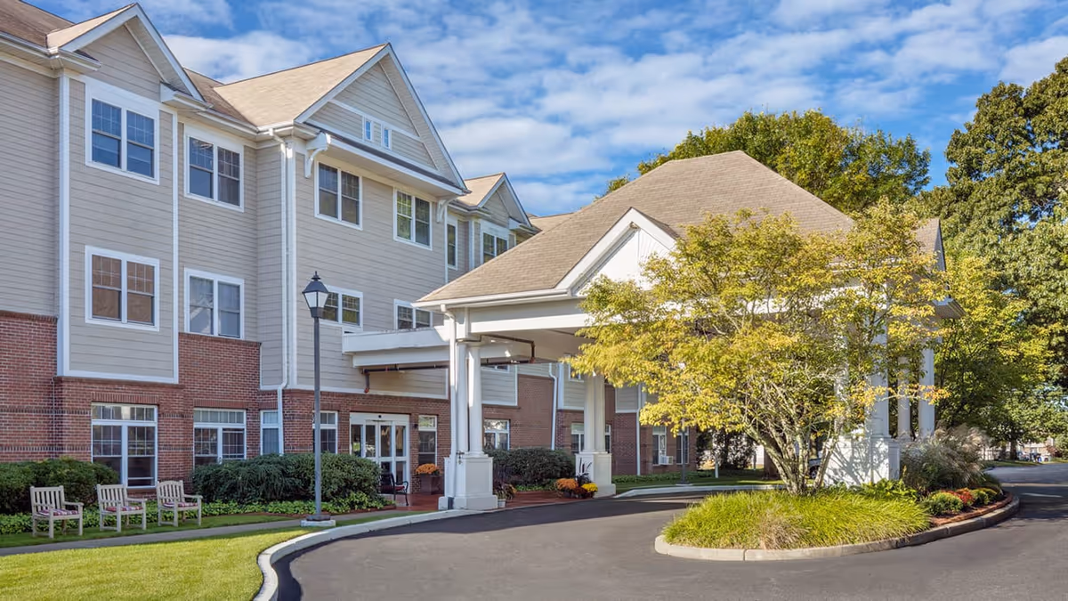 Exterior view of a senior living facility named Atria Bay Spring Village, showing a three-story building with beige siding and red brick accents. The entrance features a covered driveway with white columns, surrounded by landscaped greenery and a tree. Several wooden chairs are placed on the lawn near the building.