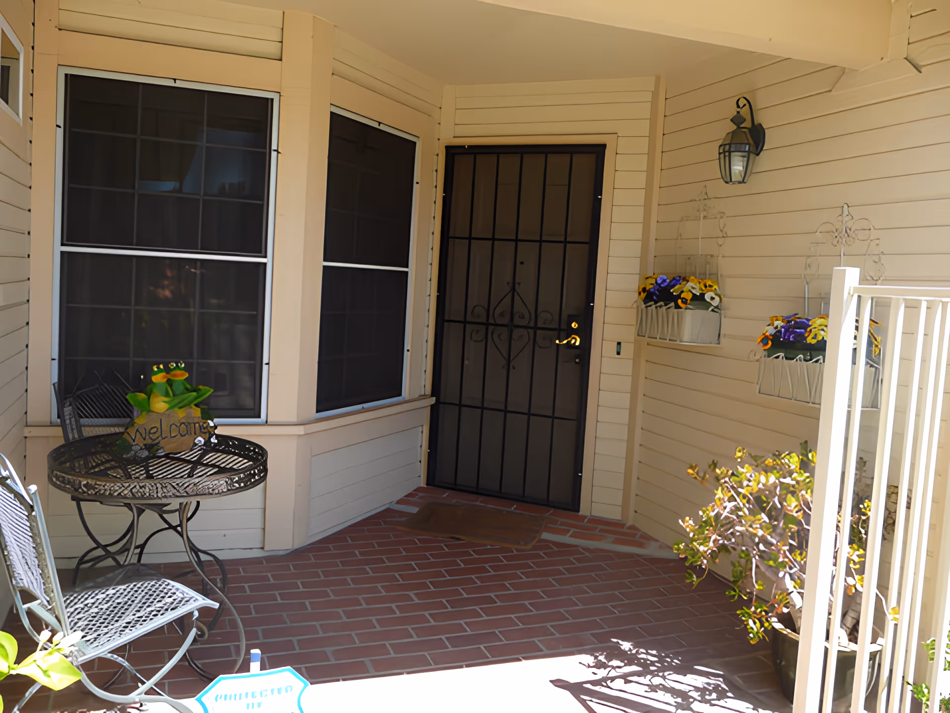Covered front porch with a brick floor, black security door and bay windows, a small metal table and chairs, potted plants and hanging flower boxes.