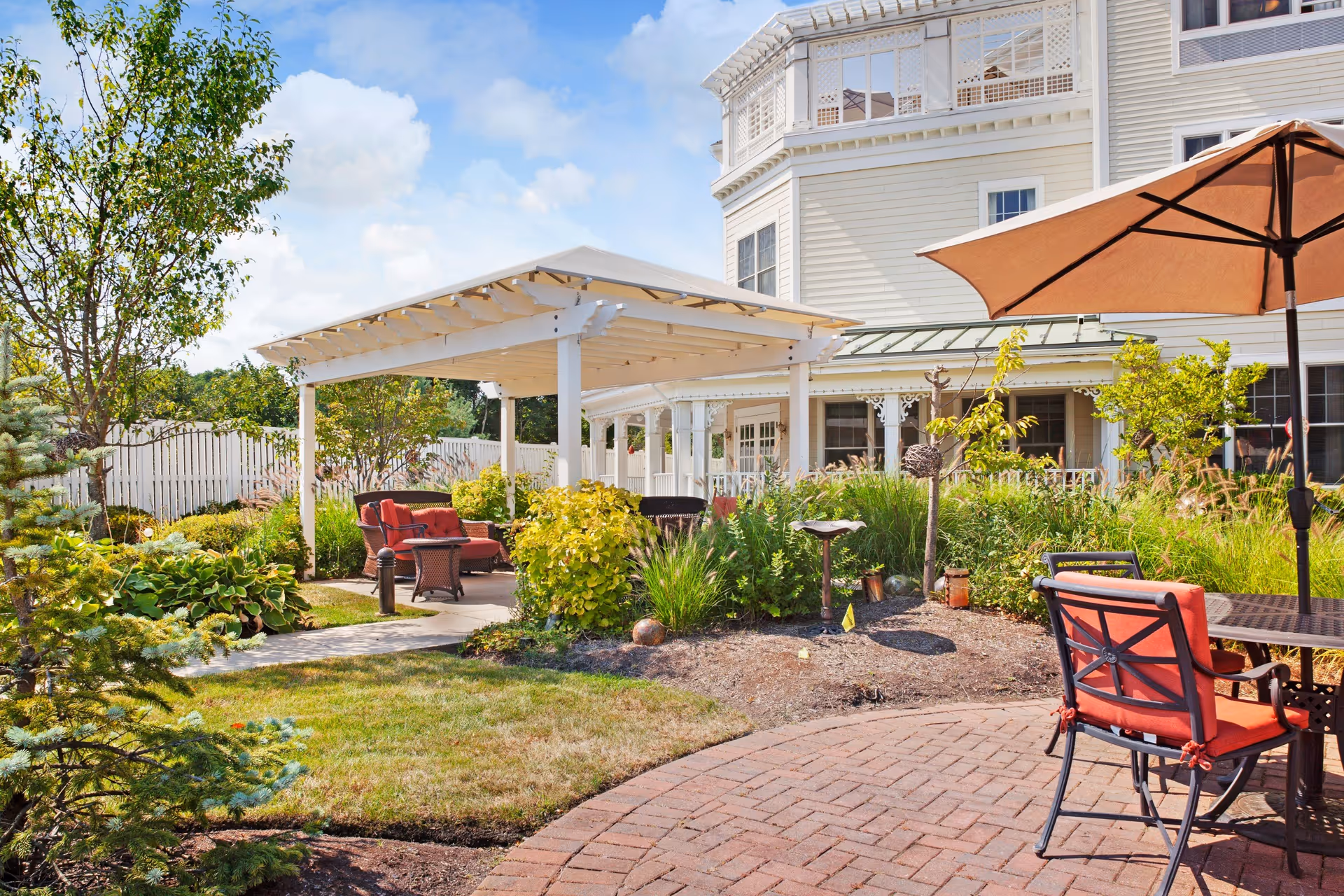 Outdoor patio area at Sunrise of Braintree featuring a white pergola with cushioned seating underneath, surrounded by green shrubs and plants. There is a brick-paved area with a table and orange cushioned chairs under a large umbrella. The building exterior is light-colored with multiple windows and decorative trim.