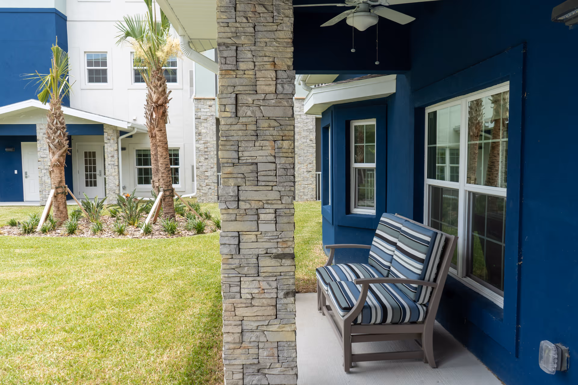 Outdoor patio area at Sunflower Springs at Trinity with a cushioned bench featuring blue and white striped upholstery, stone pillar, blue exterior walls, windows, and a ceiling fan. In the background, there are palm trees, grass, and parts of the building.
