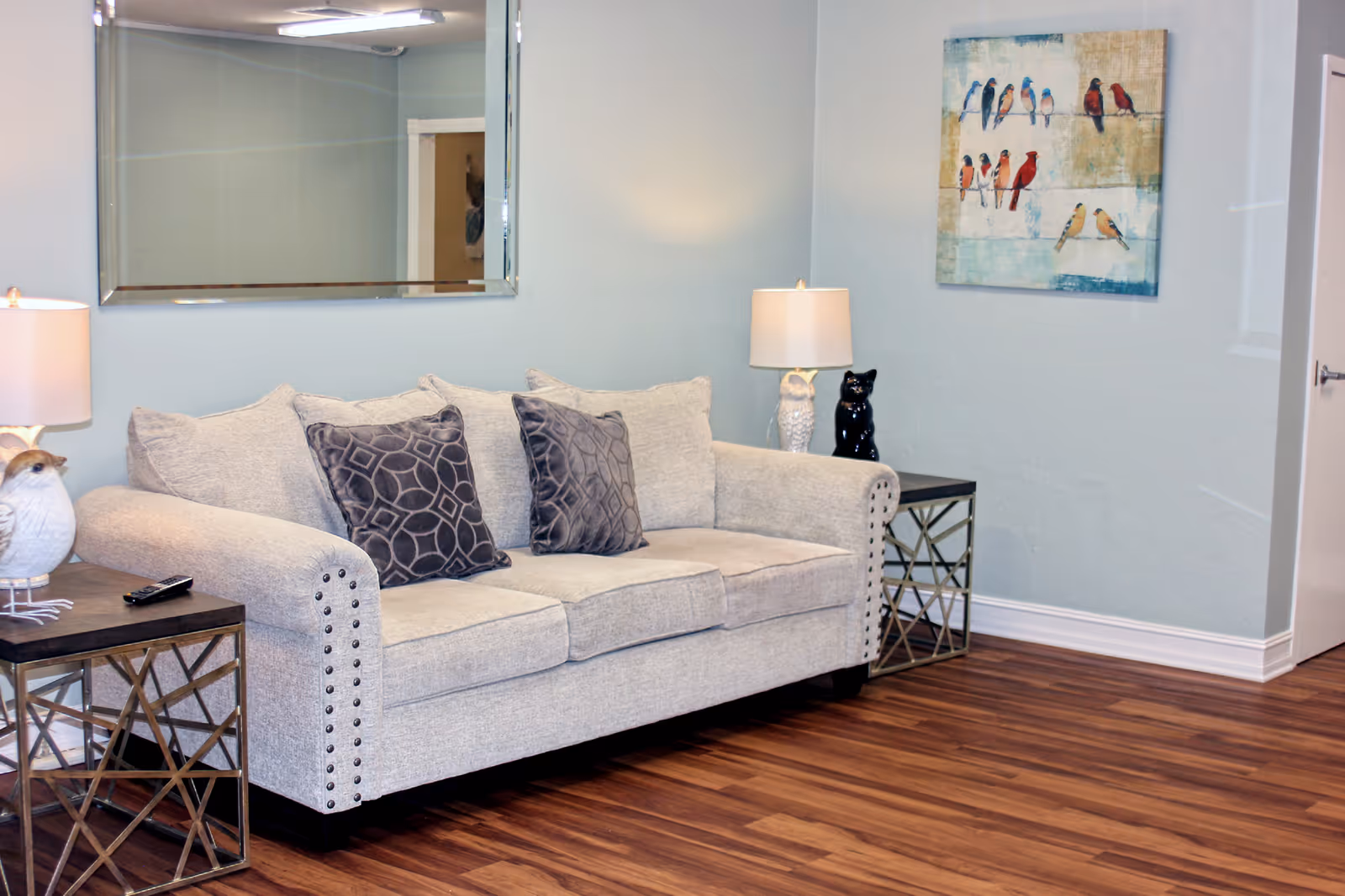 Light-gray upholstered sofa with patterned throw pillows between two end tables and lamps in a bright living room with wood floors and bird artwork on the wall.