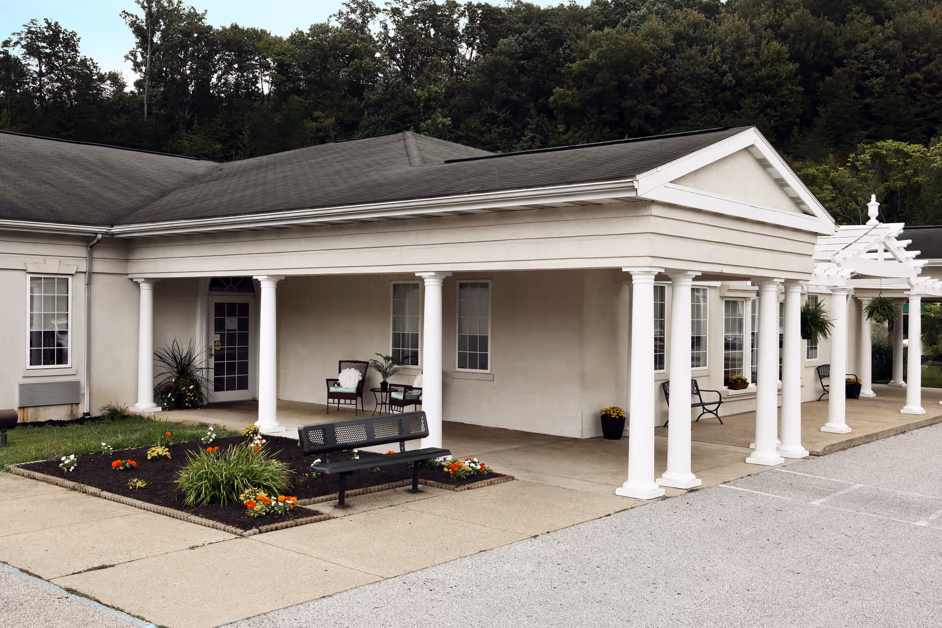 Front entrance of a single-story senior living facility with white columns, benches, potted plants, and a small flower bed.