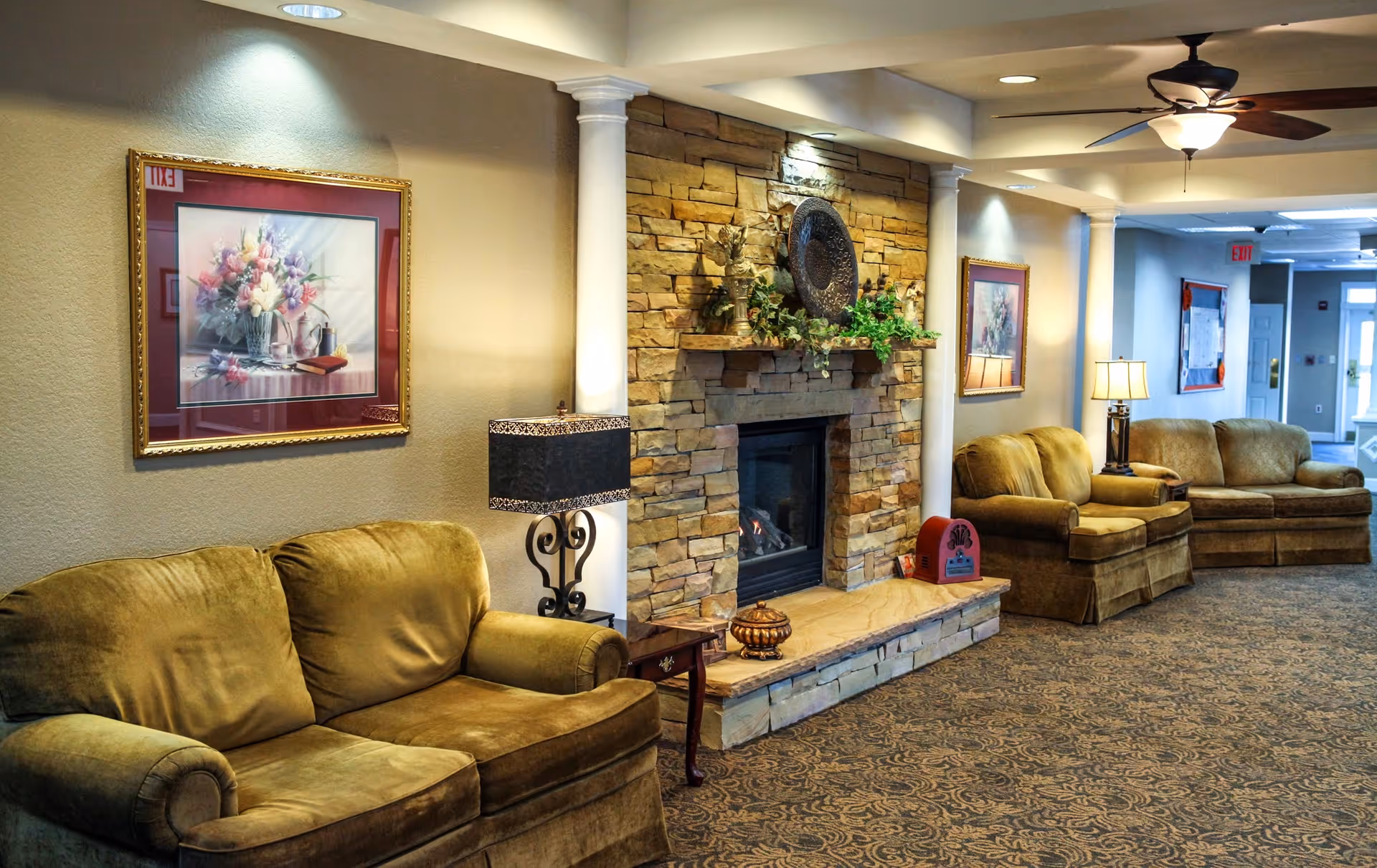 Comfortable lounge in a senior living facility featuring sofas, a stone fireplace, framed artwork, and lamps.