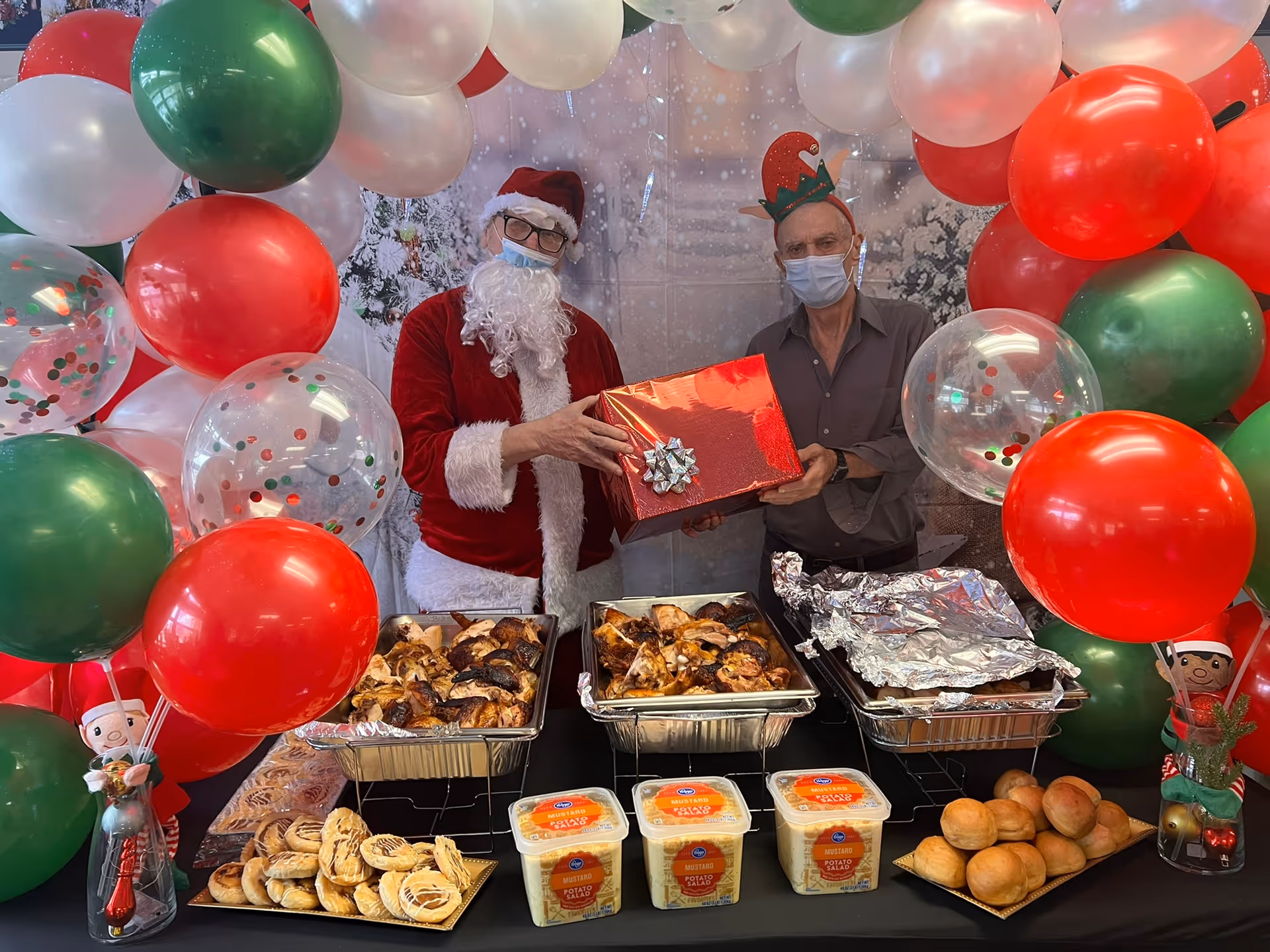Two men wearing face masks and festive holiday hats stand behind a table filled with trays of cooked chicken, rolls, pastries, and containers of potato salad. One man is dressed as Santa Claus holding a red gift box with a silver bow, while the other man wears an elf hat. The scene is decorated with red, green, white, and clear balloons with confetti inside, creating a festive holiday atmosphere.