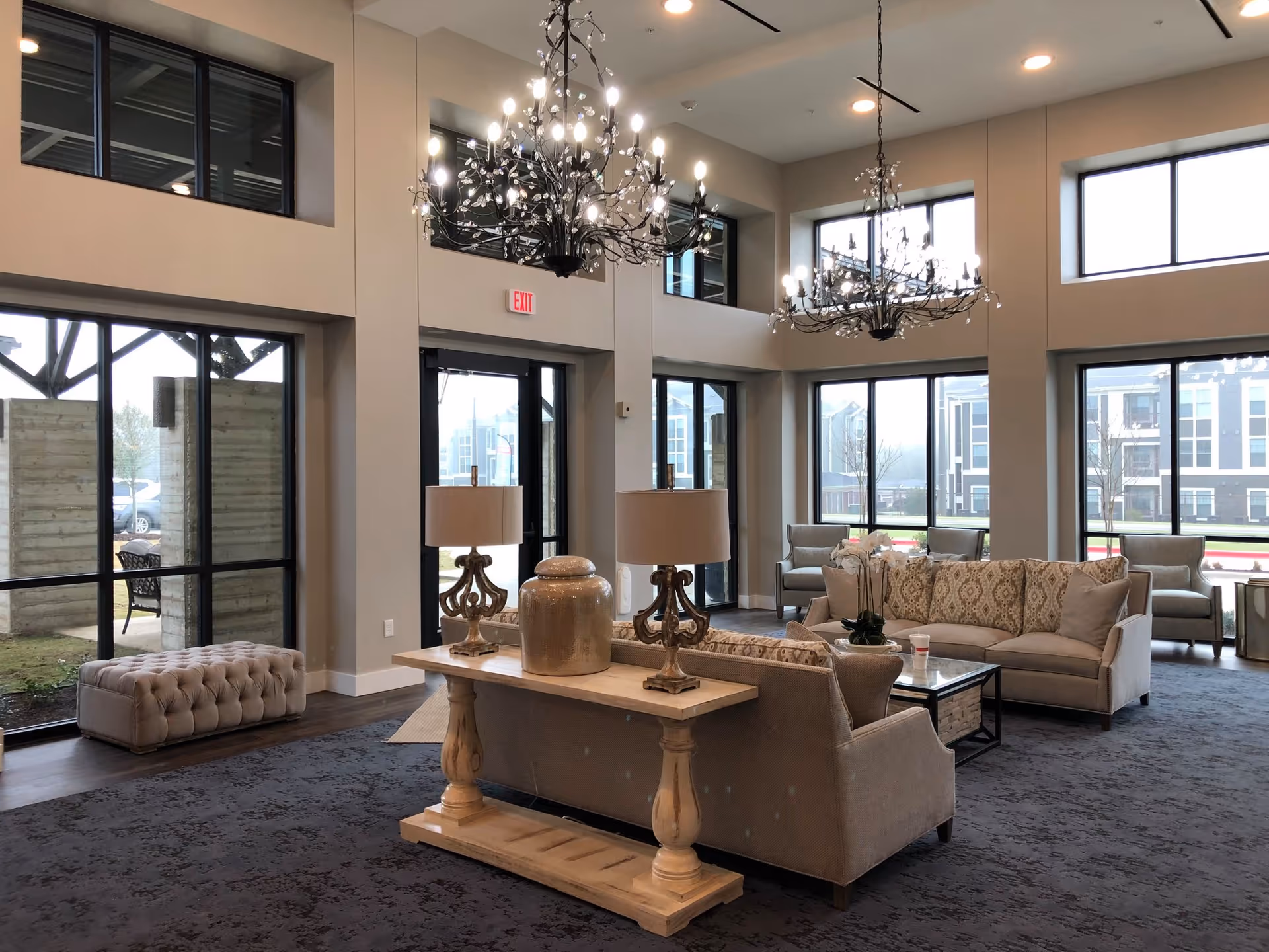 A spacious and well-lit living room area with large windows allowing natural light to fill the space. The room features two chandeliers hanging from a high ceiling, a beige sofa set with patterned cushions, a wooden table with two decorative lamps and a large vase, and a tufted bench near the window. Outside the windows, parts of a building and some greenery are visible.