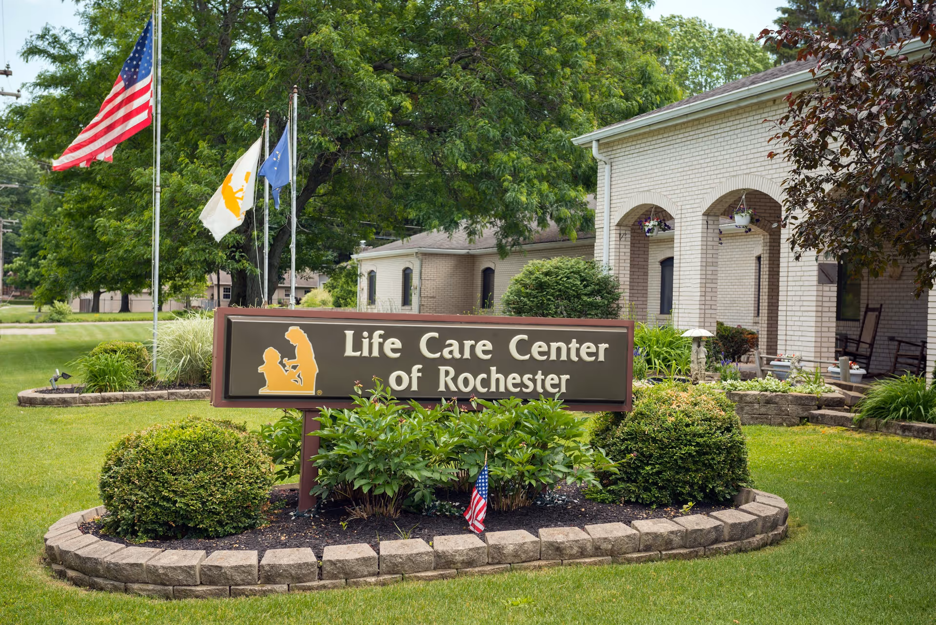 Outdoor view of the Life Care Center of Rochester sign surrounded by trimmed bushes and greenery, with three flagpoles displaying the American flag and other flags, and a light-colored brick building with a covered porch and rocking chairs in the background.