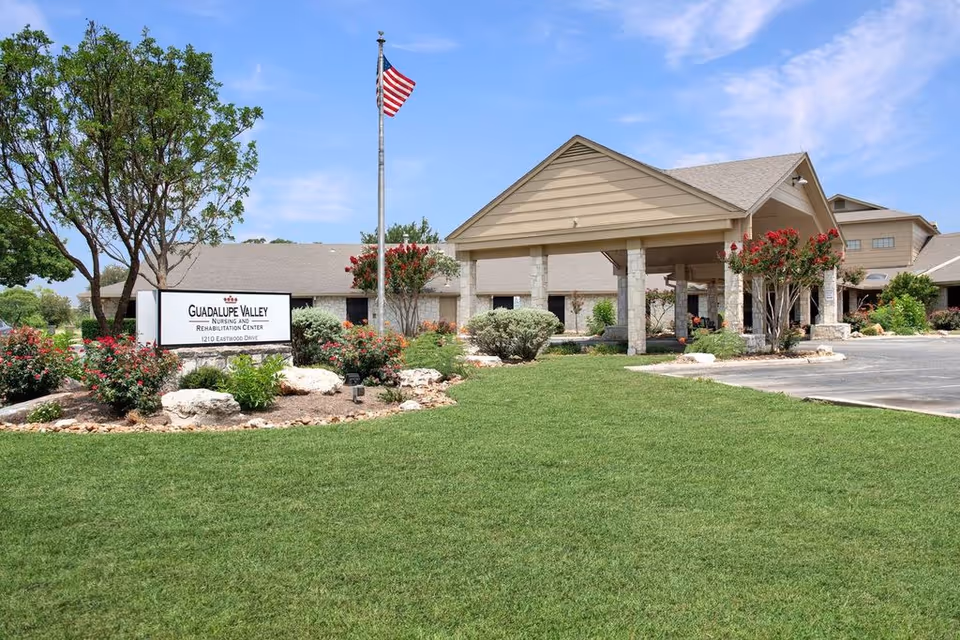 Exterior view of Guadalupe Valley Nursing and Rehabilitation Center showing a well-maintained lawn, landscaped garden with bushes and flowers, an American flag on a flagpole, and the building entrance with a covered driveway under a clear blue sky.