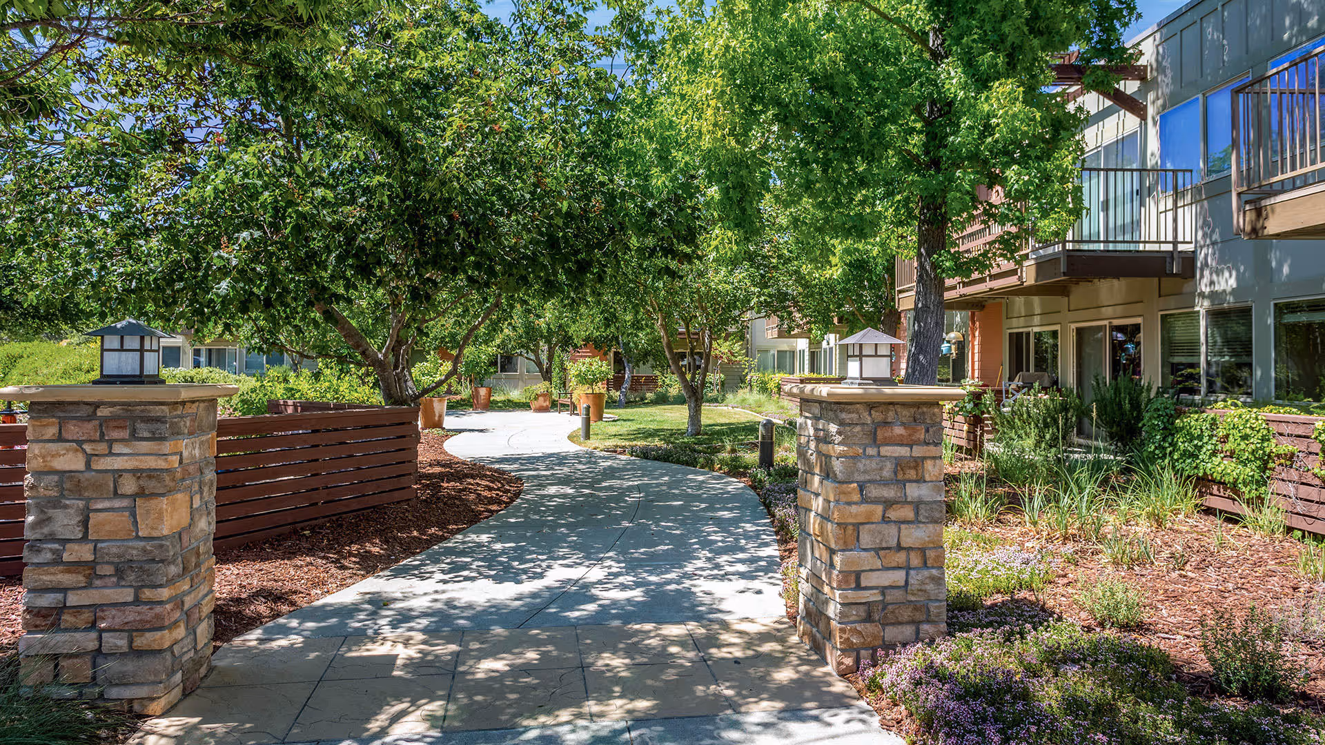 A sunny outdoor garden pathway at Atria Valley View, lined with stone pillars, wooden fences, green trees, shrubs, and flowering plants, with residential building balconies visible on the right side.
