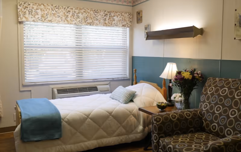 A cozy bedroom in a senior living facility featuring a single bed with a white quilted blanket and a blue throw at the foot. A striped pillow rests on the bed. Next to the bed is a wooden nightstand with a table lamp, a vase of colorful flowers, and a small decorative bowl. A patterned armchair with circular designs is positioned nearby. The room has a large window with white blinds and a floral valance, and an air conditioning unit below the window.