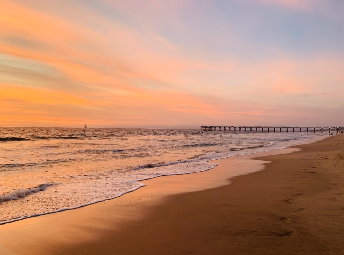 A serene beach scene at sunset with gentle waves washing onto the sandy shore. A long pier extends into the ocean on the right side, and a sailboat is visible on the horizon under a colorful sky with shades of orange, pink, and purple.