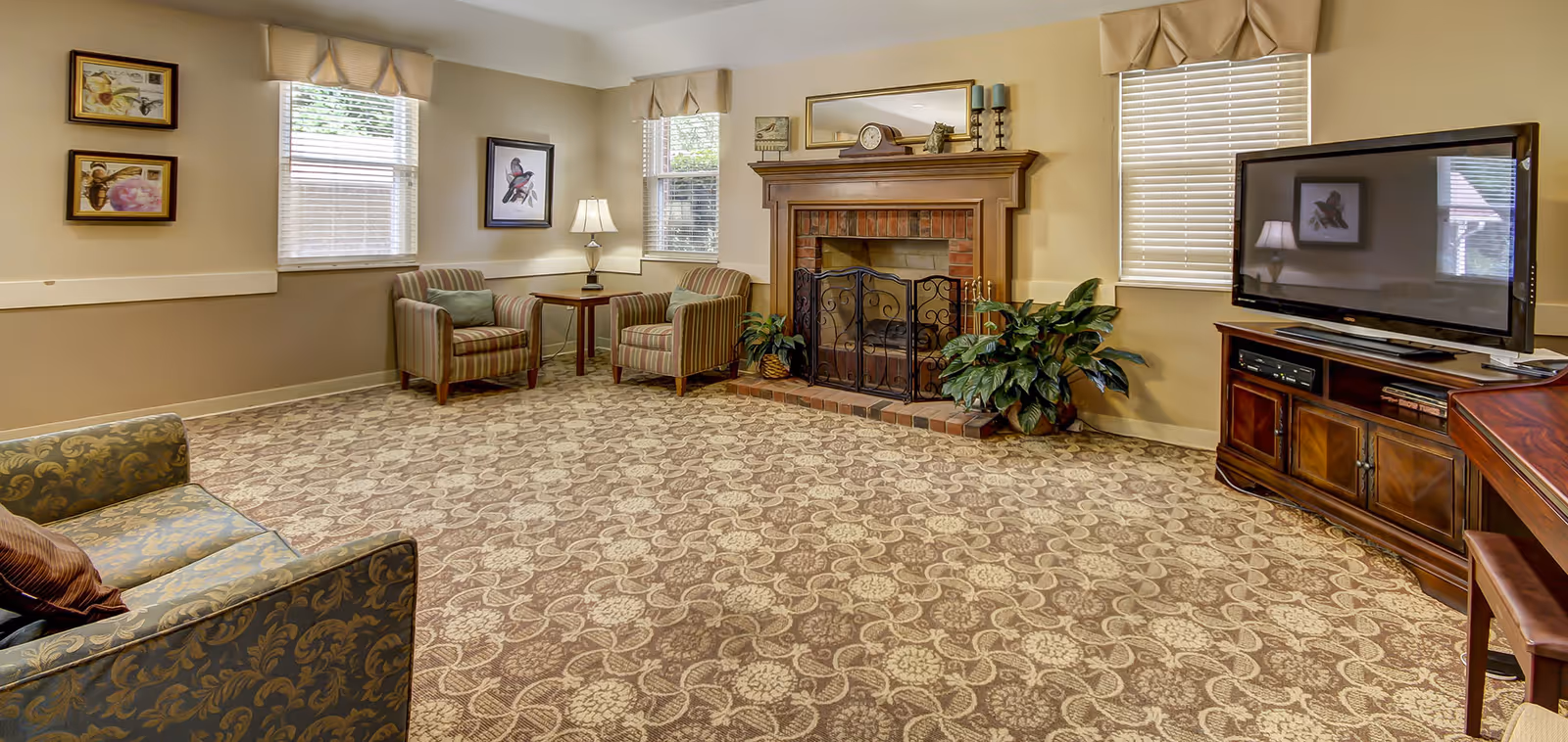 A cozy living room with patterned carpet, two striped armchairs with a small wooden table and lamp between them, a brick fireplace with a wooden mantle decorated with a clock, candles, and a small statue, a large flat-screen TV on a wooden cabinet, and a sofa with patterned upholstery. The room has three windows with blinds and beige valances, and framed artwork on the walls.