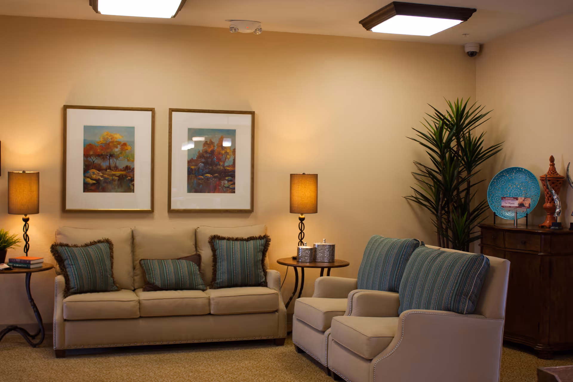 A cozy living room area with a beige sofa and two beige armchairs, all adorned with striped green and brown cushions. Two framed landscape paintings hang on the wall above the sofa. There are two side tables with lamps on either side of the sofa, and a wooden cabinet with decorative items and a tall green plant in the corner.