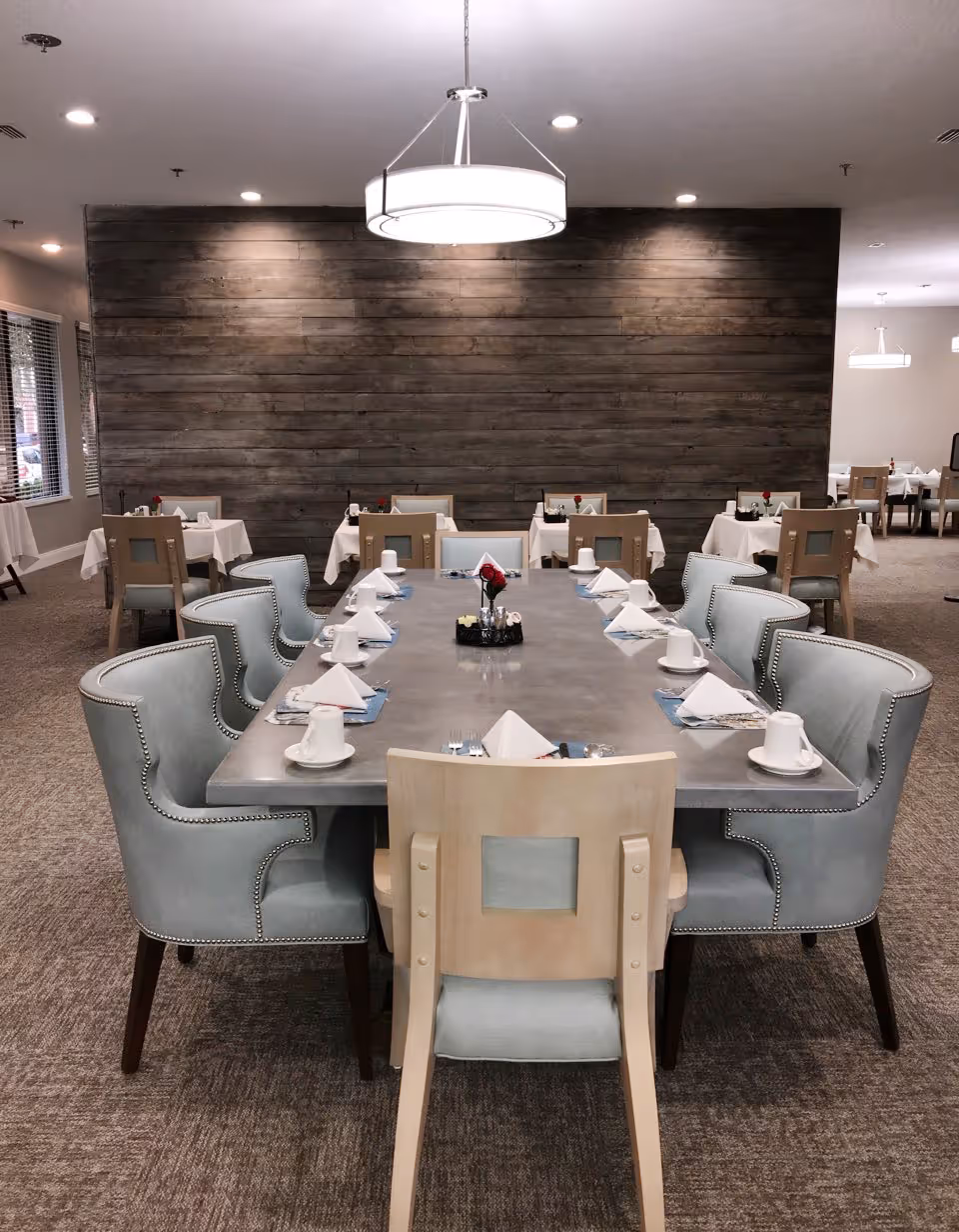 A dining room with a large rectangular table set for a meal, surrounded by light blue upholstered chairs and wooden chairs. The table is set with white napkins, cups, and silverware. In the background, there are smaller tables also set for dining, with a wooden accent wall and modern ceiling lights.