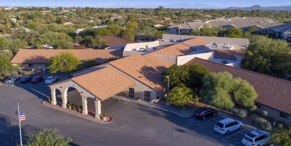 Aerial view of Mountain View Care Center showing a single-story building with a terracotta tiled roof, surrounded by trees and parked cars. The building has an arched entrance canopy and is located in a suburban area with mountains visible in the background.