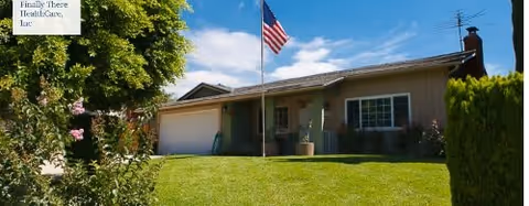 Single-story residential building with a well-maintained green lawn in front, an American flag on a flagpole, and trees and bushes surrounding the property under a clear blue sky.