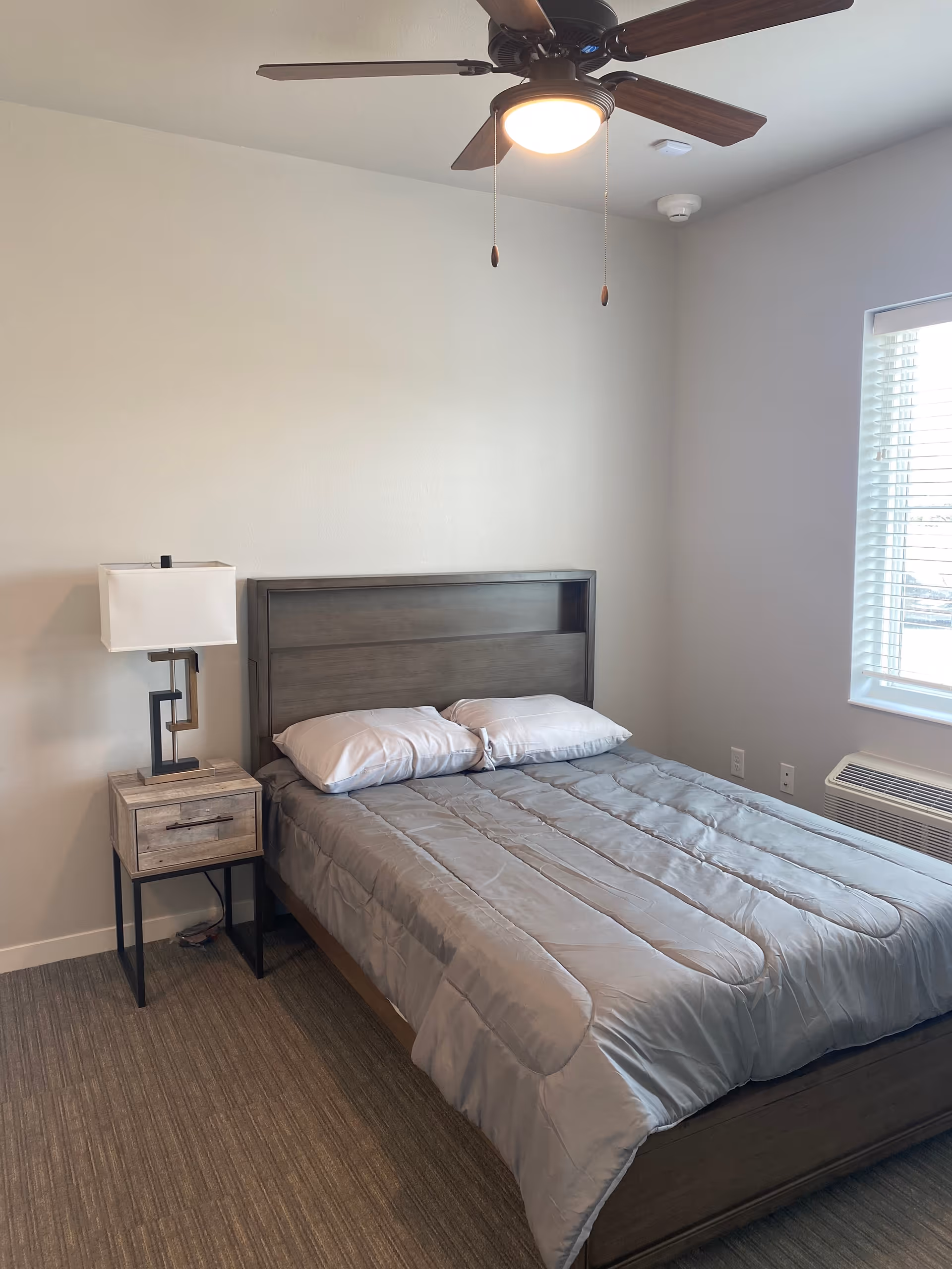 A simple bedroom with a bed made with gray bedding and two pillows, a wooden headboard, a nightstand with a modern lamp, a ceiling fan with light, a window with blinds, and a wall-mounted air conditioning unit.