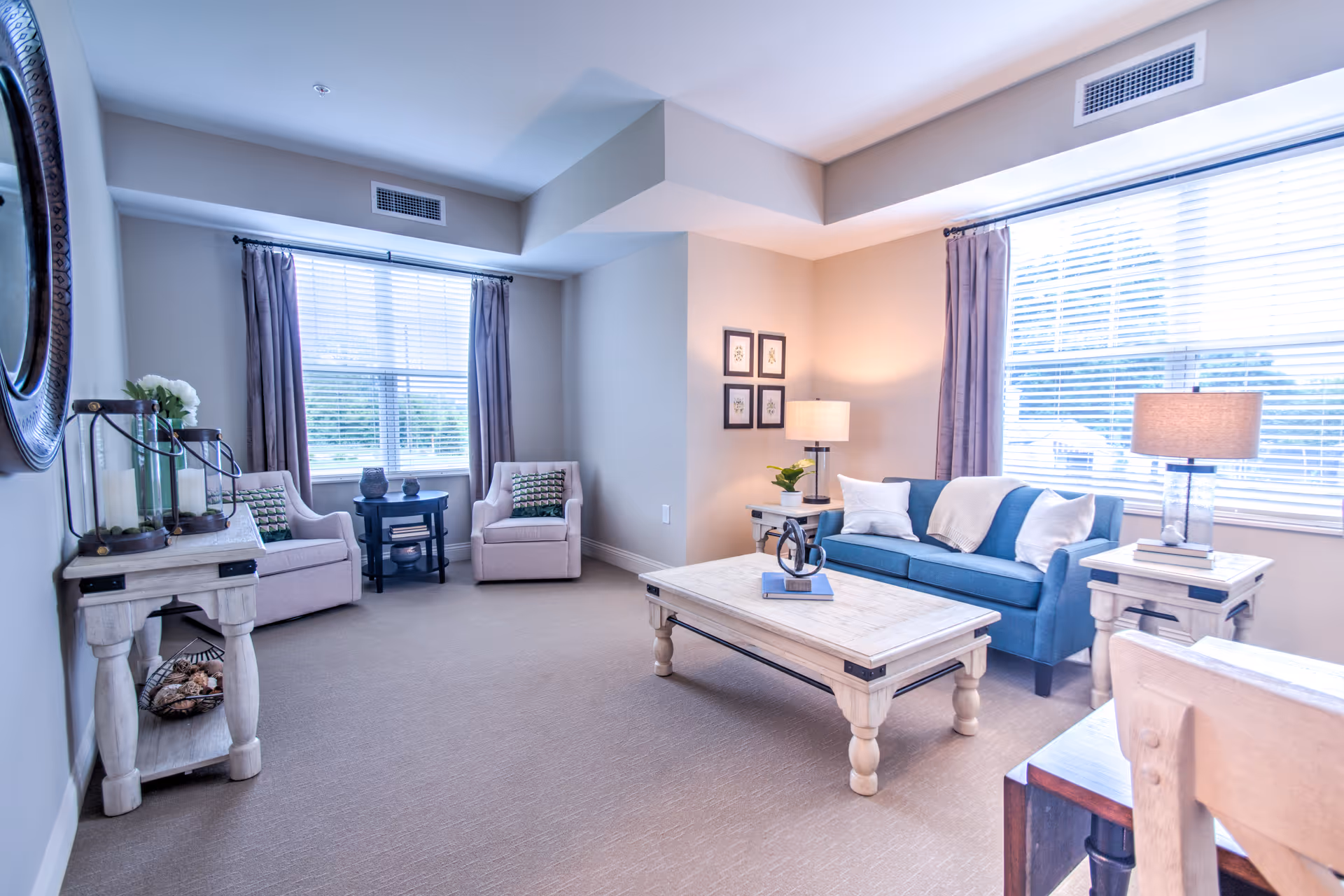 A bright and spacious living room with beige walls and carpet. The room features a blue sofa with white pillows and a throw blanket, two beige armchairs with patterned cushions, a large wooden coffee table, two side tables with lamps, and a console table with decorative items. Large windows with blinds and purple curtains allow natural light to fill the space.