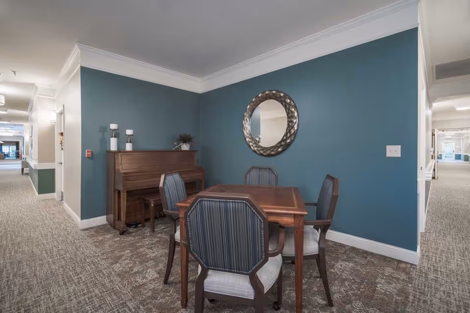 A cozy corner in a senior living facility featuring a wooden table with four upholstered chairs arranged around it. Behind the table is an upright piano with two decorative candle holders and a small plant on top. A round, textured mirror hangs on the teal-colored wall above the piano. The area is carpeted and located in a hallway with light-colored walls and ceiling molding.