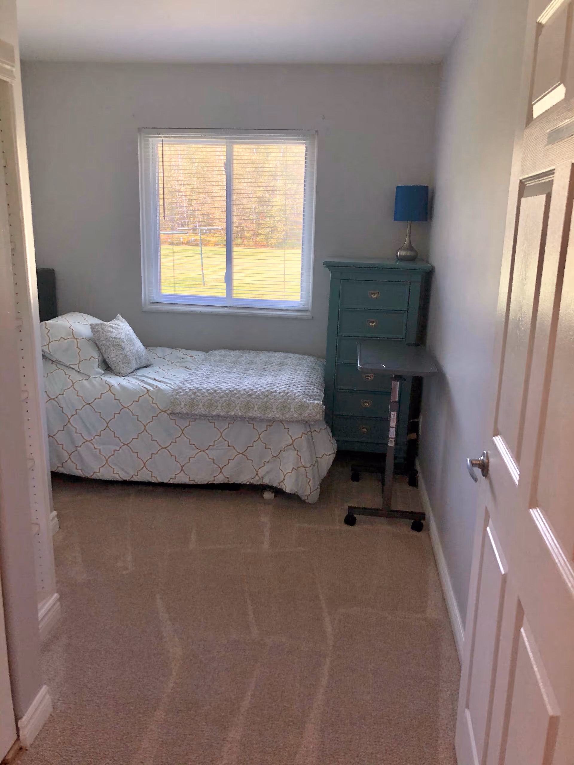 A small bedroom with a single bed covered in a white and light blue patterned comforter and pillow. There is a window with blinds letting in natural light, a teal chest of drawers with a blue lamp on top, and a small black rolling table next to the chest. The room has beige carpet and white walls.