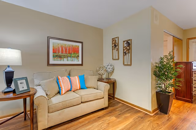 A cozy living room area with a beige loveseat adorned with three colorful throw pillows in blue and orange stripes. To the left of the loveseat is a wooden side table with a lamp and a framed photo. Above the loveseat hangs a framed painting of a forest with red and orange trees. On the right side, there is another small wooden side table with decorative items and a tall green potted plant near an open doorway leading to another room. The floor is wooden and the walls are painted a light beige color.