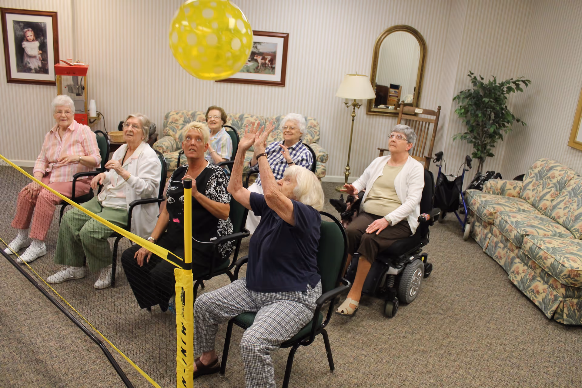 A group of elderly women seated in chairs in a common room, playing a seated balloon volleyball game with a yellow balloon and a small net. The room has patterned couches, a standing lamp, framed pictures on the wall, and a potted plant.