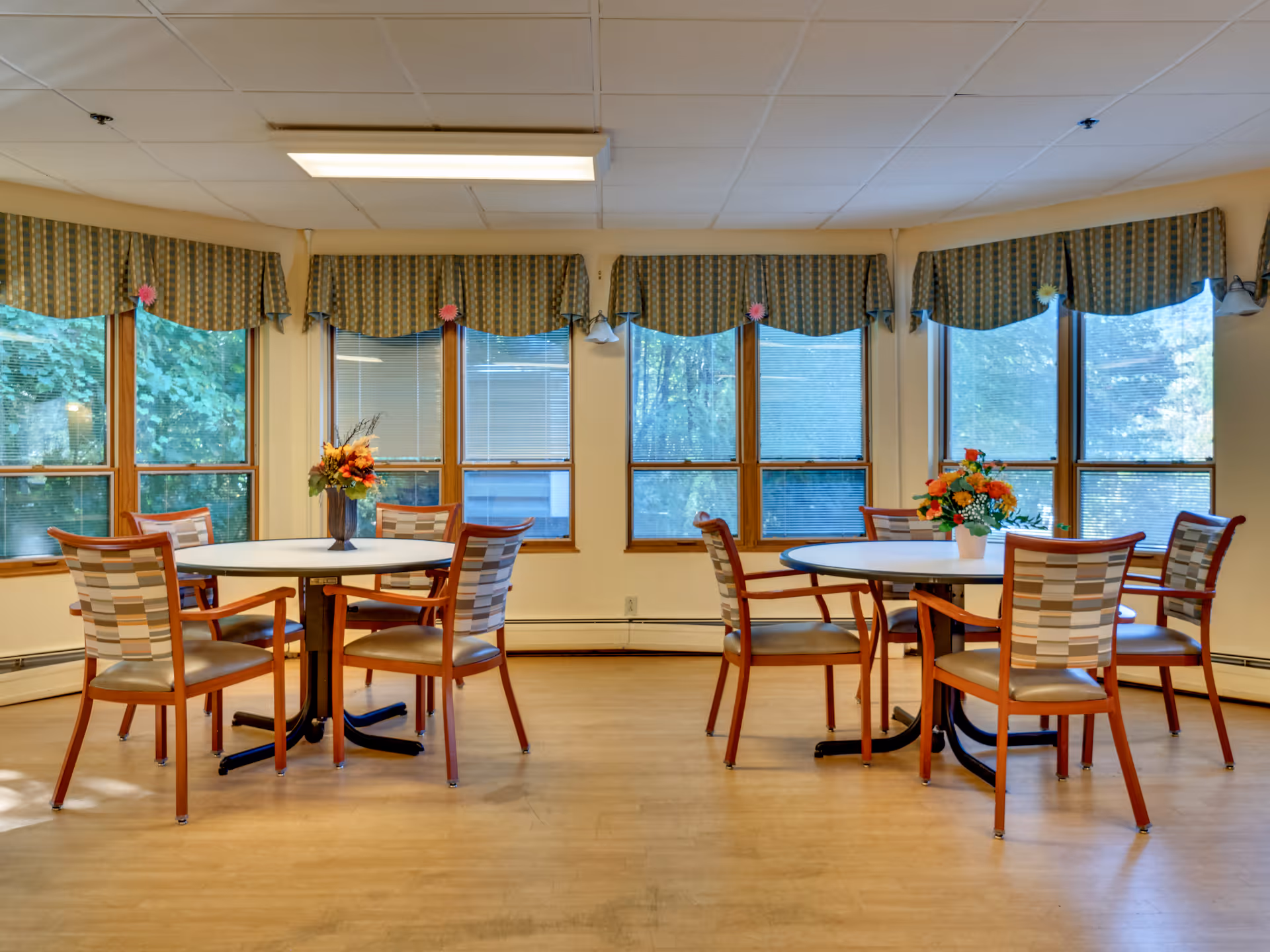 A bright room with large windows covered by patterned valances, featuring two round tables each surrounded by four wooden chairs with cushioned seats. Each table has a vase with colorful flowers as a centerpiece. The floor is wooden, and the walls are light-colored.