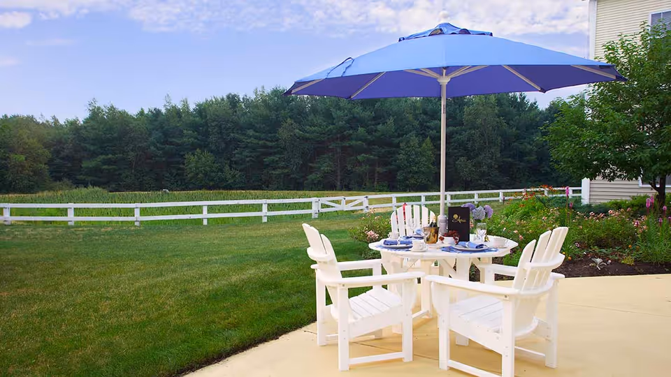 Patio with white Adirondack chairs around a table under a blue umbrella overlooking a grassy yard and white fence.