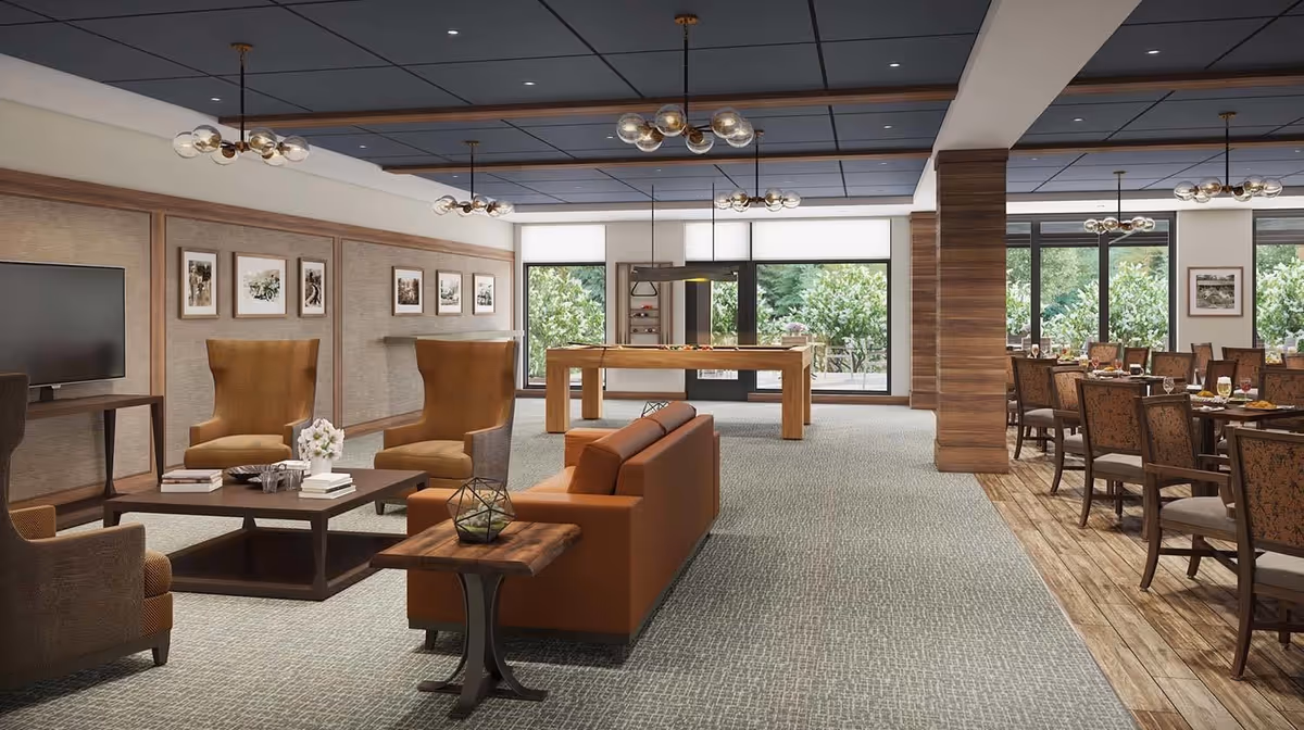 A spacious and well-lit common area in a senior living facility featuring a seating area with a brown leather sofa, two high-back armchairs, a coffee table with books and flowers, a pool table in the background, and a dining area with multiple tables and chairs set for a meal. Large windows provide natural light and views of greenery outside.