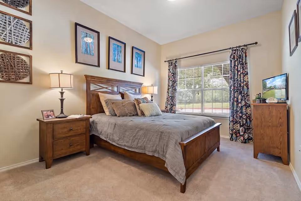 A cozy bedroom with a wooden bed frame and a gray comforter, several pillows, two wooden nightstands each with a lamp, framed artwork on the walls, a large window with floral curtains letting in natural light, and a wooden dresser with a TV on top.