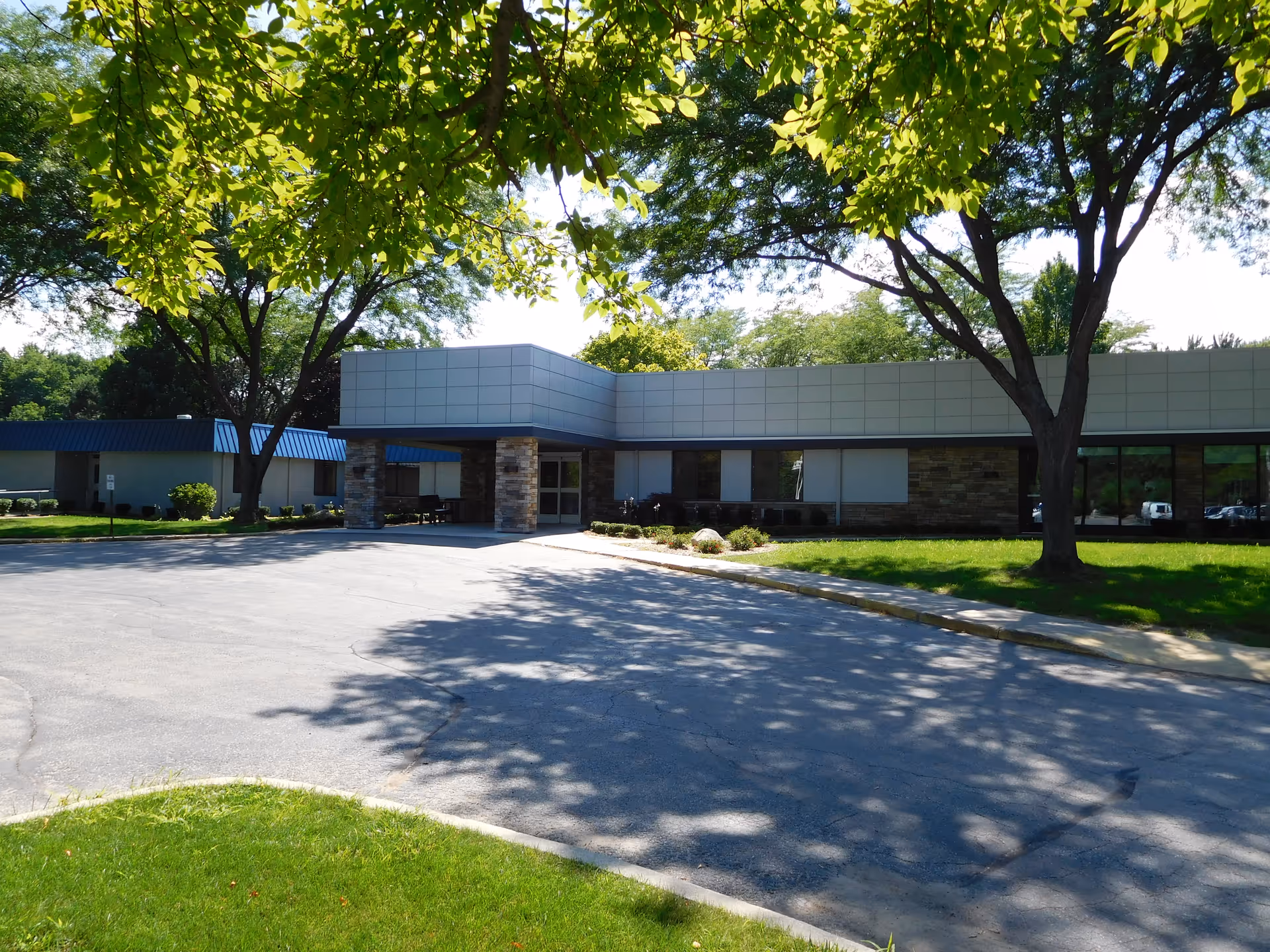 Exterior view of a single-story building with a covered entrance supported by stone pillars, surrounded by green grass and trees casting shadows on the driveway in front.