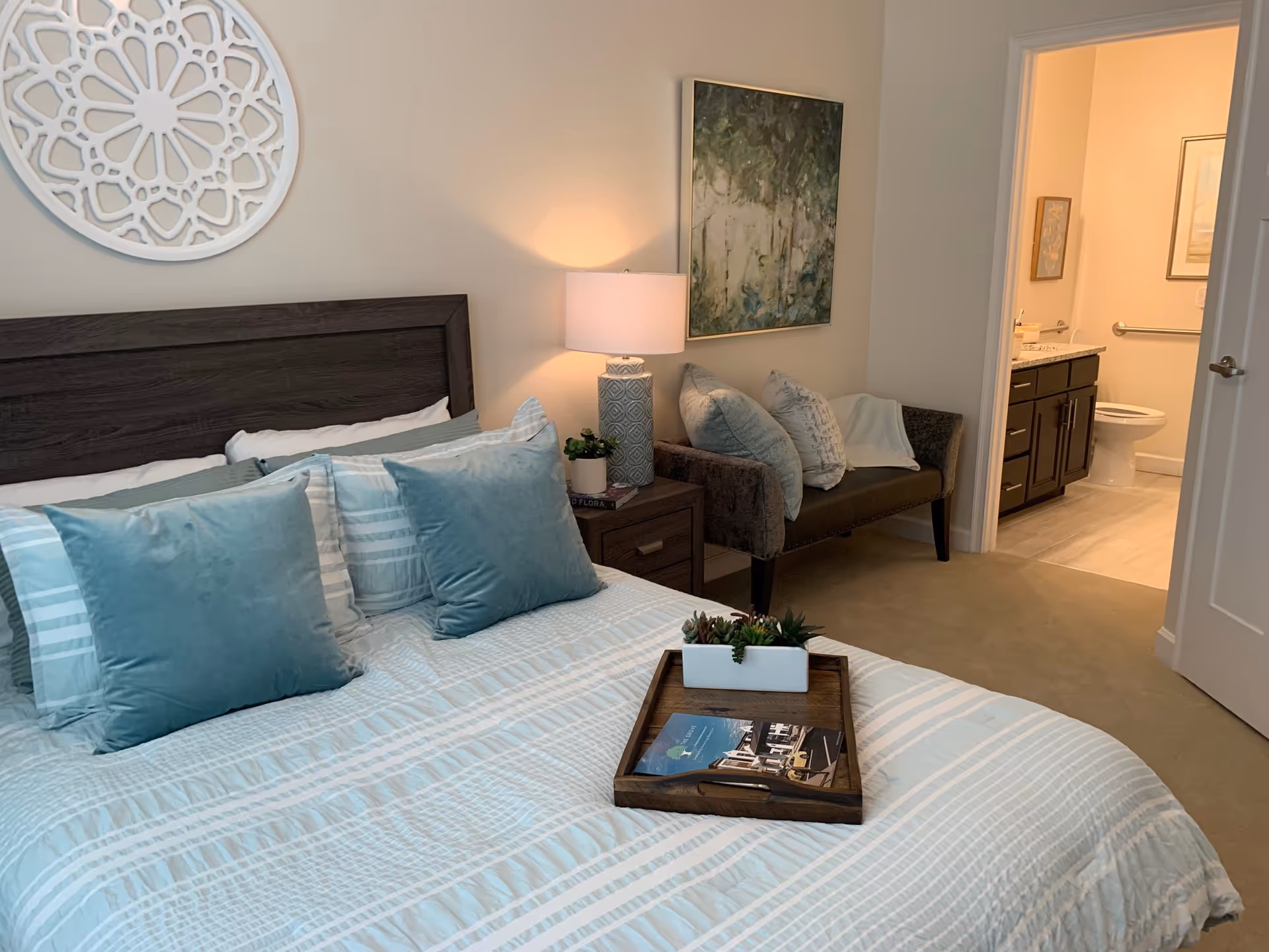 Well-decorated bedroom with a made bed and tray, bedside table with lamp, bench seating, and an open door showing an adjoining bathroom.