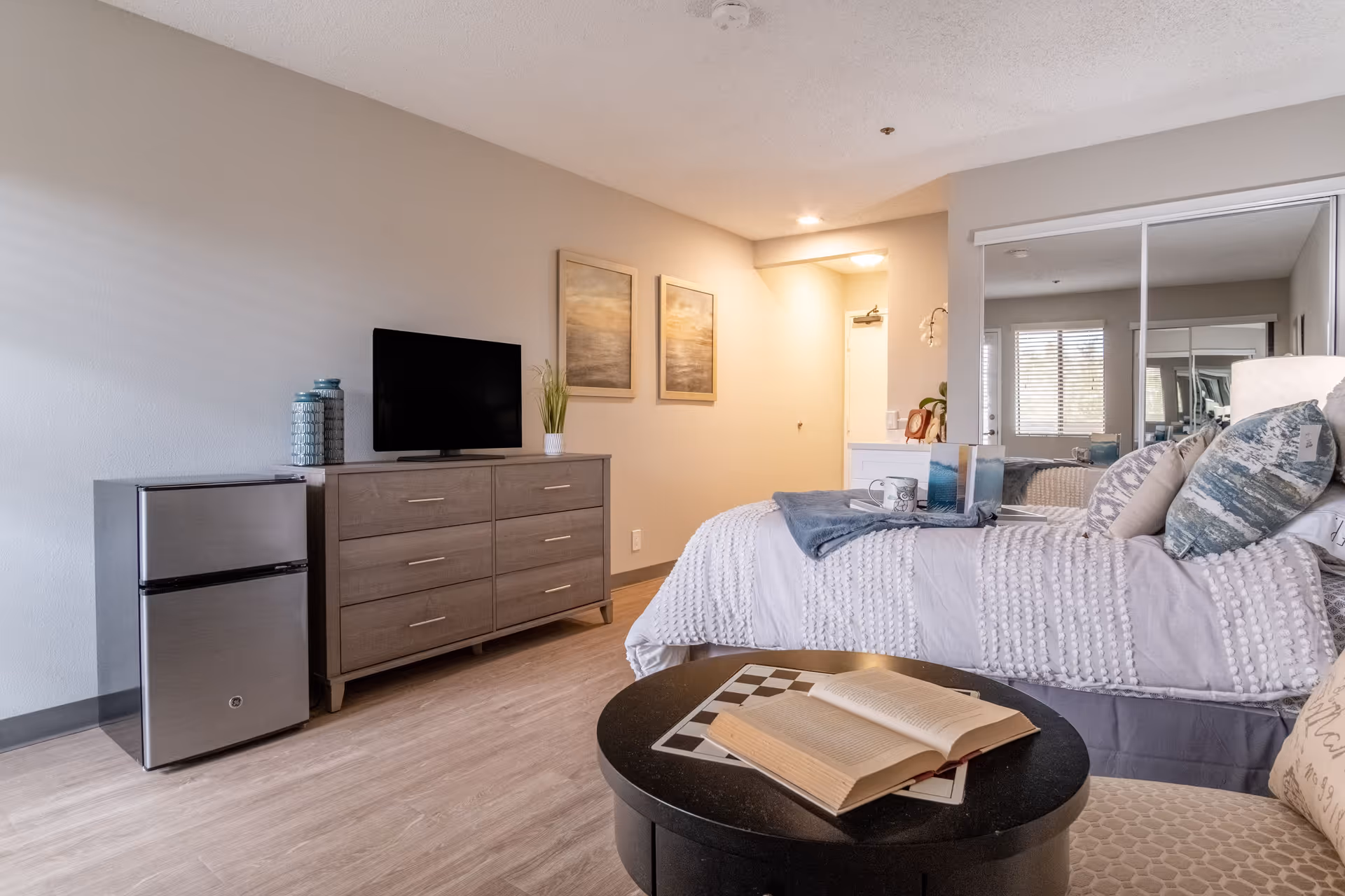 A cozy bedroom in an assisted living facility featuring a bed with white textured bedding and multiple pillows, a round black table with an open book on top, a dresser with a flat-screen TV and decorative vases, and a small stainless steel refrigerator. The room has light-colored walls, wood flooring, two framed paintings on the wall, and a mirrored closet door reflecting a window with blinds.