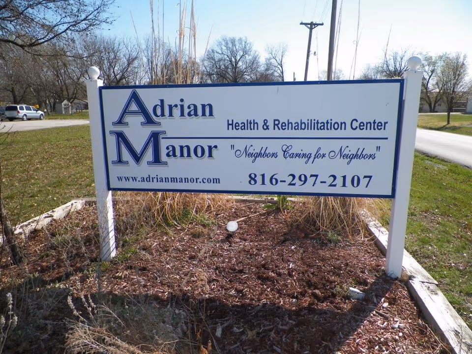 Outdoor sign for Adrian Manor Health & Rehabilitation Center with the slogan 'Neighbors Caring for Neighbors', a website URL, and a phone number, situated on a grassy area with trees and a road in the background.