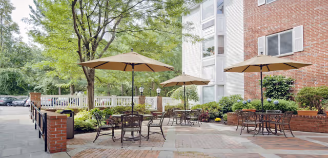 Outdoor courtyard with metal patio tables and umbrellas beside a brick-and-siding building and surrounding trees.