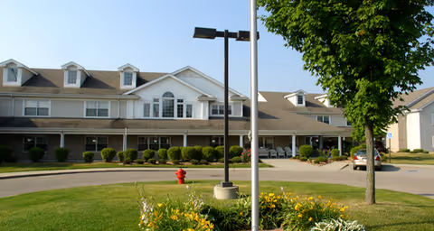 Exterior view of a large senior living facility building with multiple dormer windows, a covered entrance, landscaped bushes, a tree, and a flower bed with yellow flowers in the foreground. A red fire hydrant and a street lamp are also visible near the entrance.