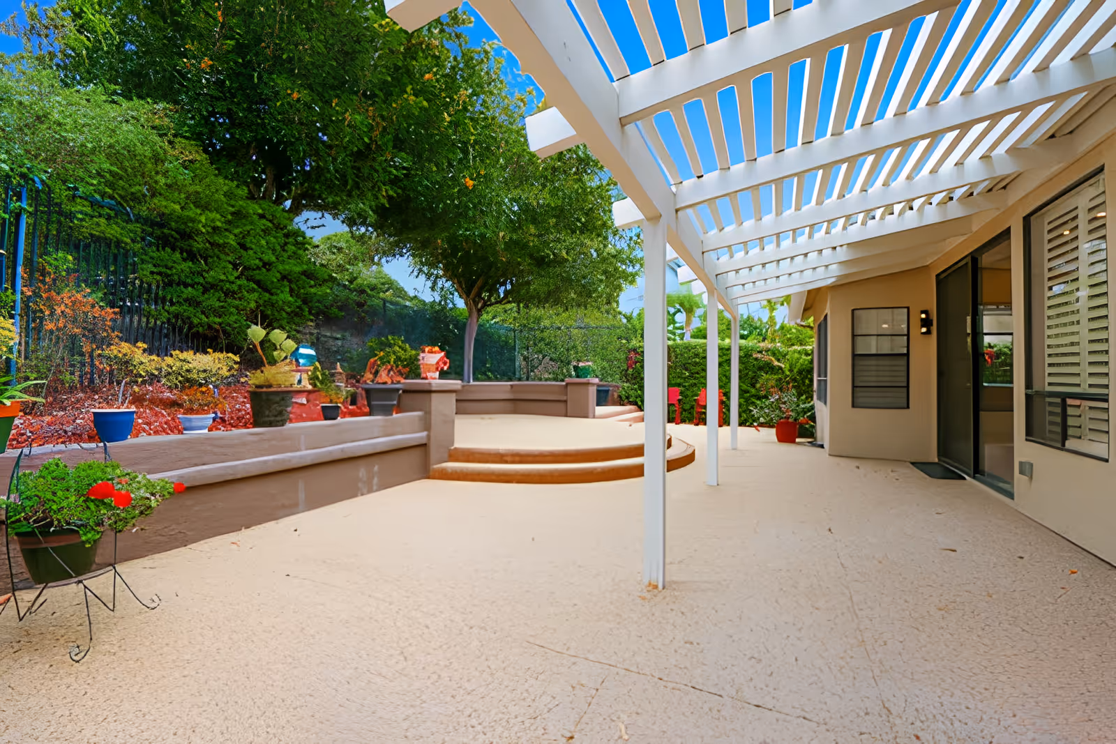 Outdoor patio area with a white pergola overhead, beige textured flooring, potted plants along a low wall, and a few steps leading to a raised section surrounded by greenery and trees under a clear blue sky.