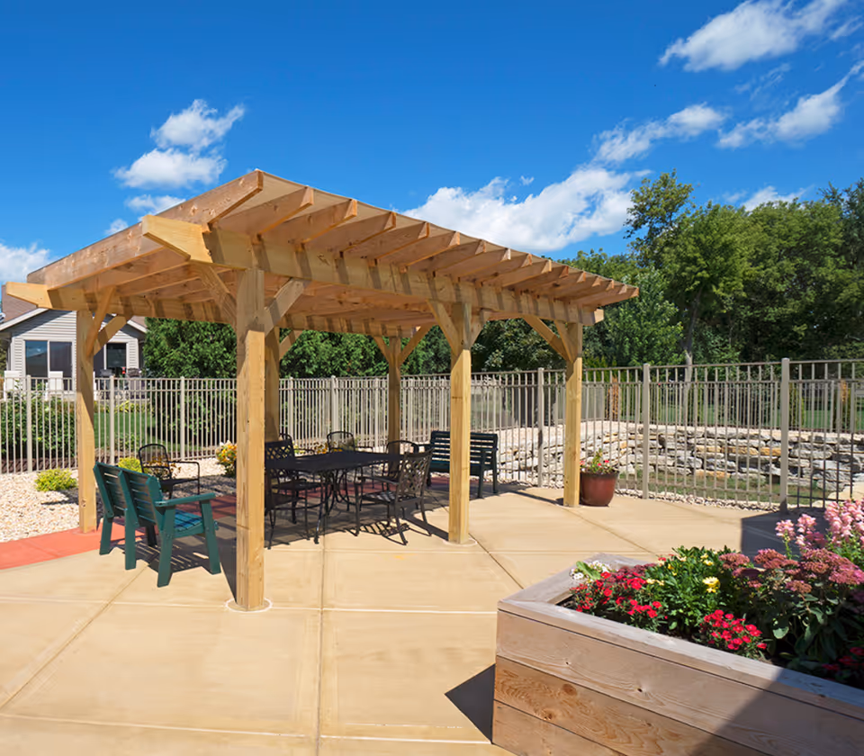 Outdoor patio area at Pine Villa Memory Care featuring a wooden pergola with seating underneath, including green benches and black metal chairs around a table. The area is surrounded by a metal fence, with a planter box filled with colorful flowers in the foreground and trees in the background under a blue sky with scattered clouds.
