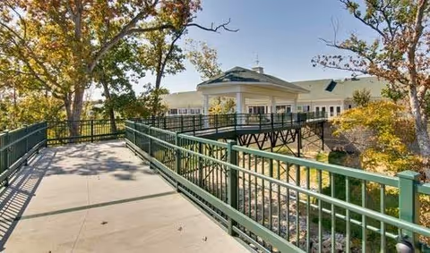 A paved outdoor walkway with green metal railings surrounded by trees with autumn foliage, leading to a building with a light-colored exterior and green roof.