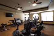 Interior view of a fitness room with exercise equipment including stationary bikes and elliptical machines. The room has large windows with blinds, ceiling fans, and a wall-mounted television.
