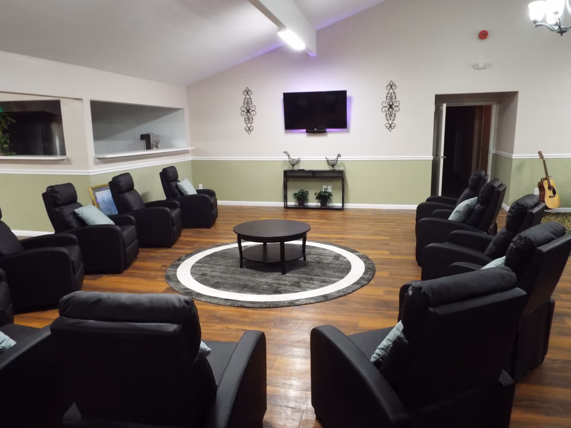 A spacious common room with black recliner chairs arranged in a circle around a round coffee table on a circular rug. The room has wood flooring, light green and white walls, a wall-mounted flat screen TV, decorative wall hangings, and a guitar resting against the wall in the corner.