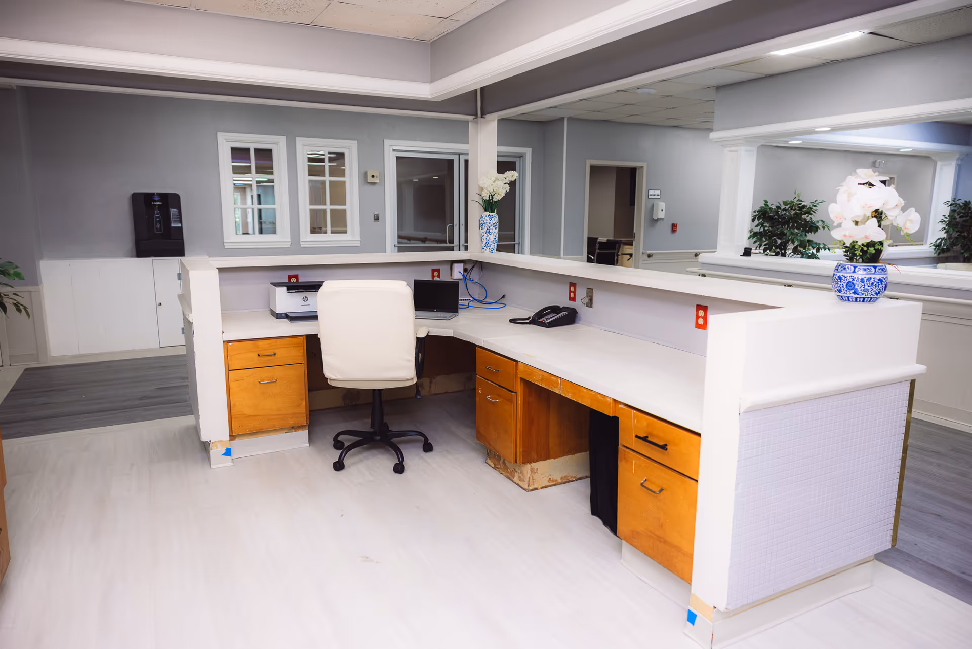 Reception desk area in a healthcare facility with a white swivel chair, wooden drawers, a printer, a telephone, and decorative vases with flowers. The walls are painted gray and white, and there are windows and doorways visible in the background.