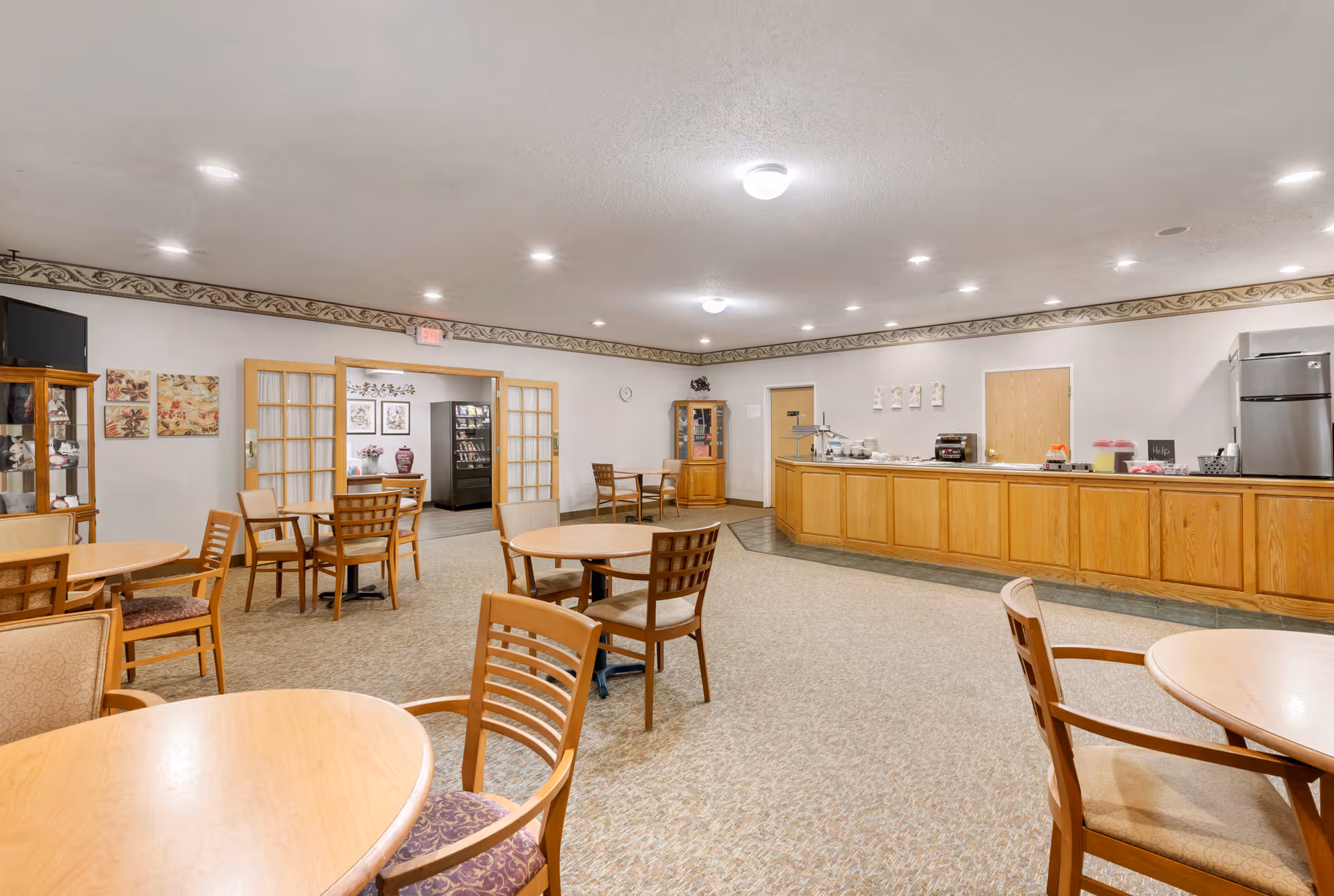 A spacious dining room with multiple round wooden tables and chairs arranged throughout. The room features a long wooden counter along one wall with a refrigerator and various items on top. The walls are decorated with framed artwork and a glass cabinet displaying decorative items. The ceiling has recessed lighting and the floor is carpeted.