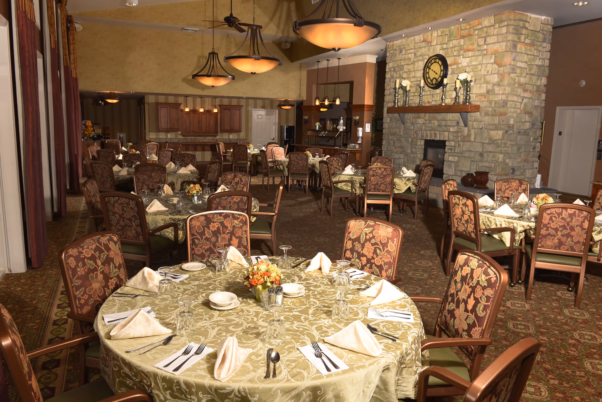 A spacious dining room in a retirement community with multiple round tables covered with patterned tablecloths, each set with napkins, plates, glasses, and silverware. The room features upholstered chairs with floral patterns, a large stone fireplace with a clock and candles on the mantel, warm lighting fixtures hanging from the ceiling, and a kitchen area in the background.