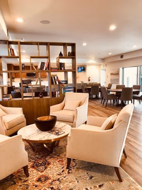 A cozy and well-lit living area in a senior living community featuring four beige upholstered armchairs arranged around a round marble coffee table with a decorative bowl on top. Behind the seating area is a wooden open bookshelf filled with books and decorative items. In the background, there are multiple dining tables with chairs, a wall-mounted TV, and large windows allowing natural light to enter the space.