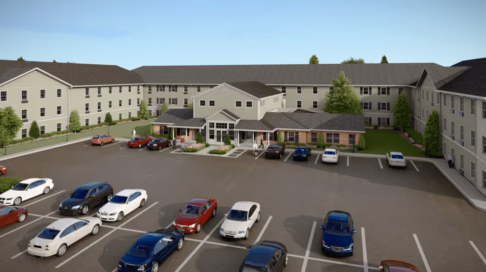 Front view of a three-story senior living facility with a central entrance, landscaped grounds, and a parking lot with cars.